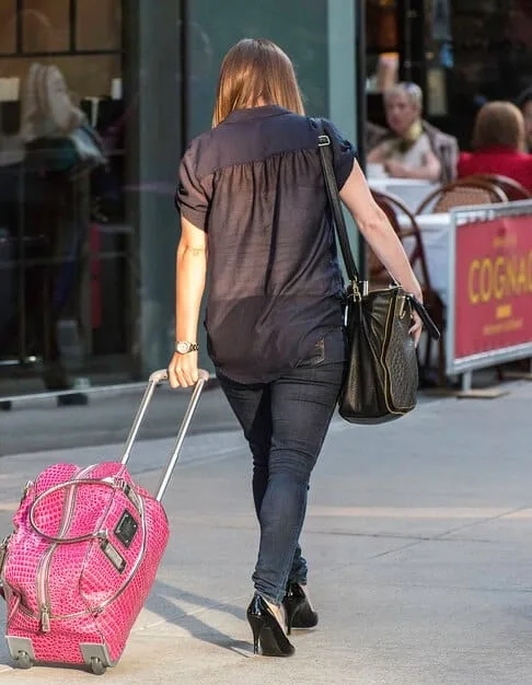 A woman confidently walking through a bustling US airport terminal, pulling a stylish carry-on suitcase behind her. She is dressed in a chic yet comfortable travel outfit, consisting of a tailored blazer, dark jeans, and fashionable sneakers.