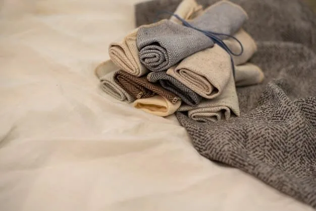 A close-up shot of a neatly arranged pile of clothing in neutral colors, including a white blouse, dark jeans, a gray sweater, and a black blazer. The clothing is folded or rolled tightly to save space, and a packing cube is visible in the background.