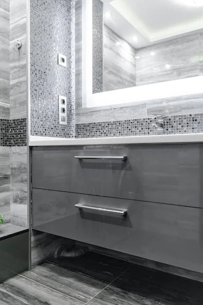 Close-up of a bathroom vanity with a faux-marble countertop, ceramic tile backsplash, and stylish brushed nickel faucet. There's a small, decorative soap dispenser and folded hand towels on the counter. The lighting is warm, highlighting the details of the materials.