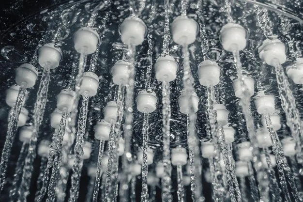 A close-up of a modern showerhead with a rainfall effect, installed in a tiled shower stall. The tiles are a light gray color, and the showerhead is chrome. Water droplets are visible, showcasing the showerhead in action.